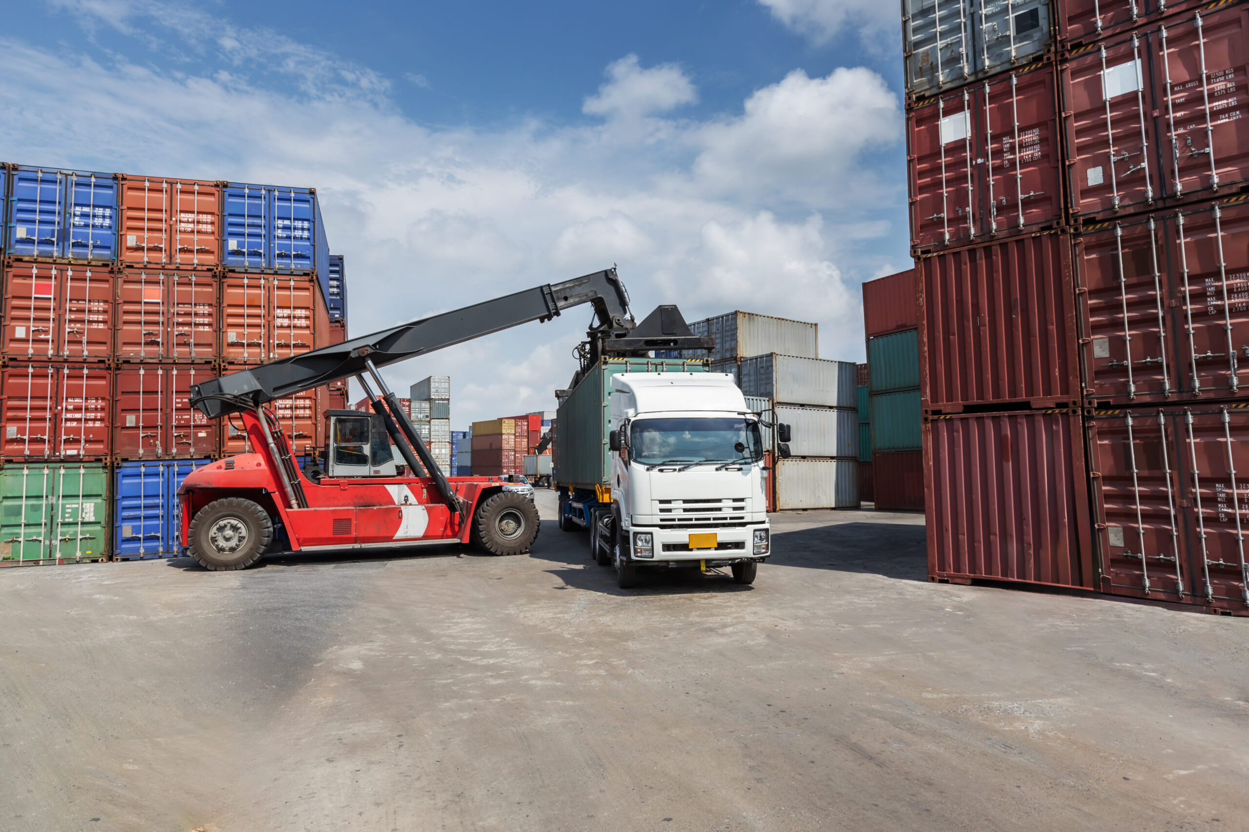 A red container forklift loads a cargo container onto a white truck in a busy shipping yard. Surrounded by stacked containers, the scene represents global logistics, trade, and freight transportation.